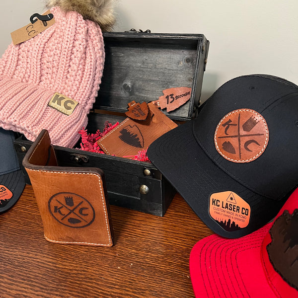 Collection of hats and a wallet with 'KC Laser Co' branding on a wooden surface.