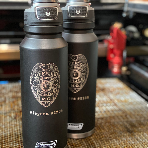 Two black Coleman water bottles with police department logo on a textured surface.